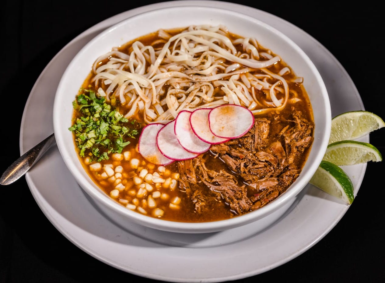 A bowl of birria ramen: noodles in red consommé with shredded birria beef, corn, sliced radish and cilantro, limes on the side