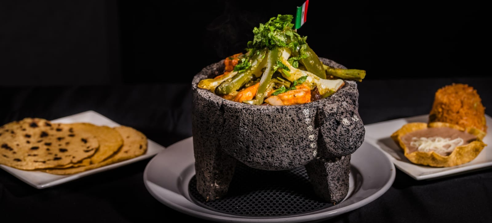 A molcajete bubbling in its stone bowl, grilled meats, cactus and cheese over the sauce