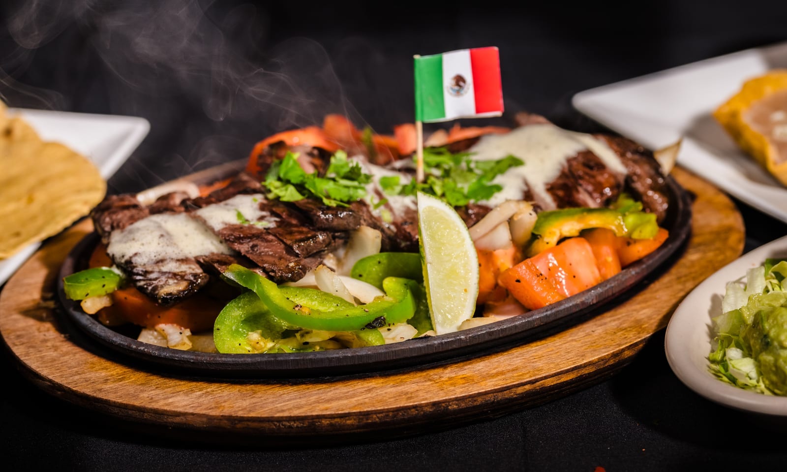 Steak fajitas on a sizzling skillet with peppers and onions, tortillas and sides alongside
