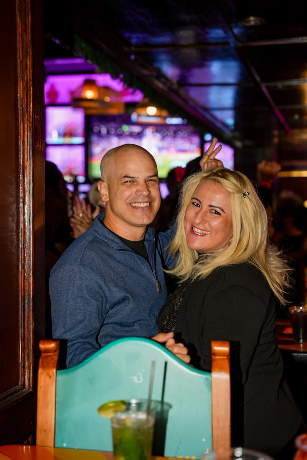 A couple smiling together at a booth, a drink on the table and the lit bar behind them
