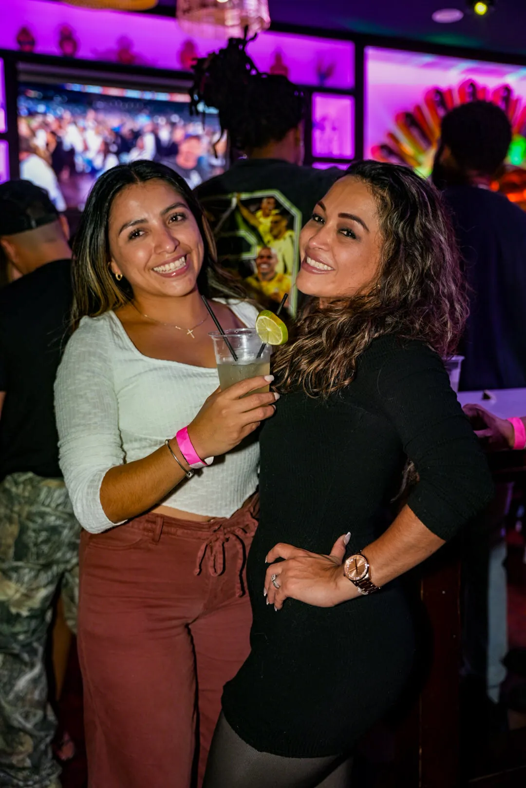 Two friends with drinks posing together at the bar under the pink shelf lights