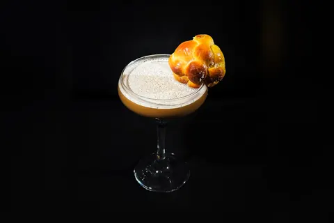 Dulce Descanso cocktail in a stemmed coupe with a sugar-dusted foam top and a mini pan de muerto on the rim, on a black background