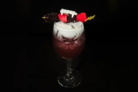 Berry Scary cocktail, deep red with a white foam top, garnished with a skewer of blackberries, raspberries and vampire fangs, on a black background