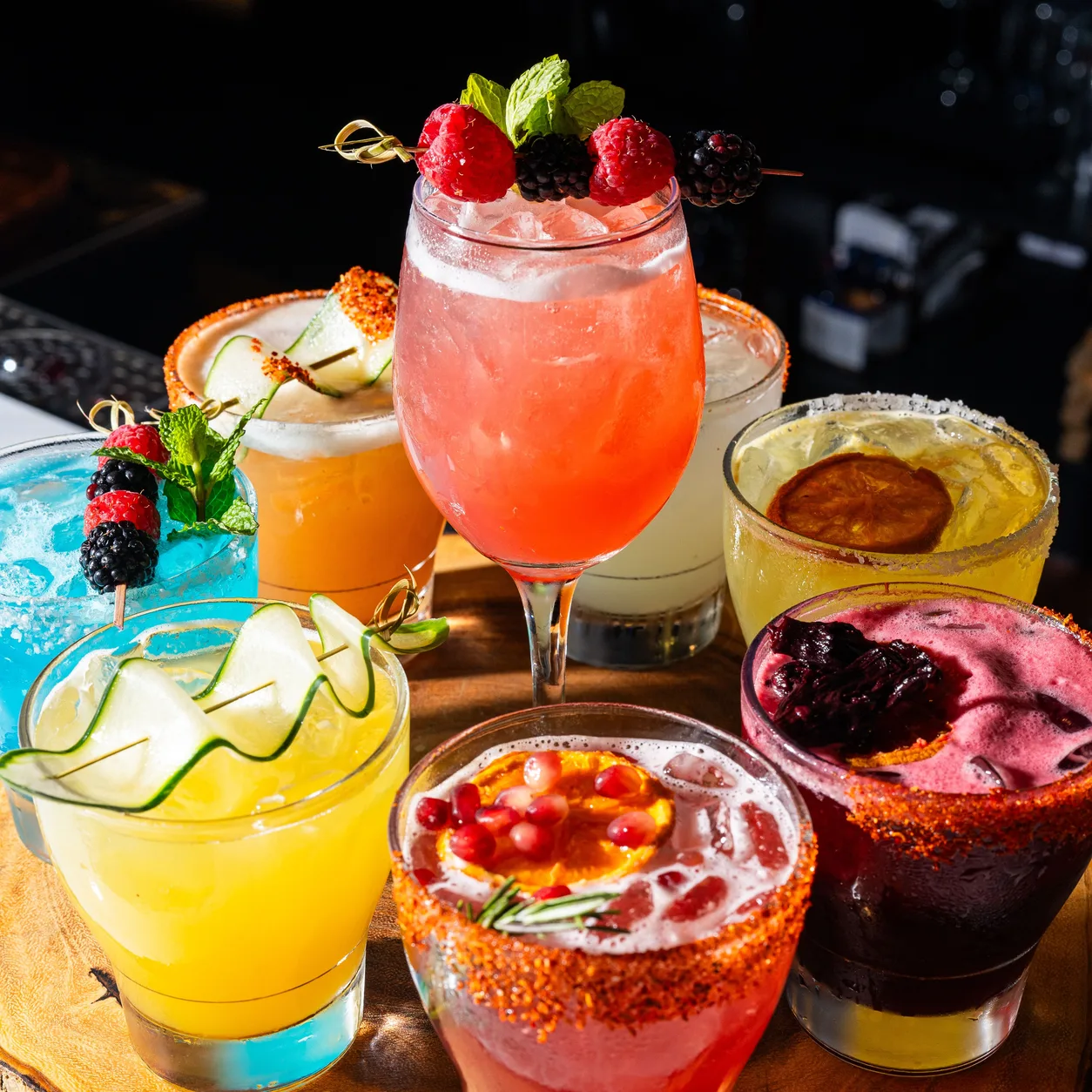 Eight handcrafted cocktails on a wooden board: a berry-topped spritz, chile-rimmed margaritas, a blue drink and a jamaica margarita