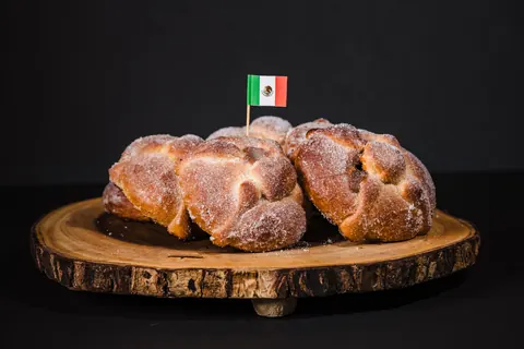 Three sugar dusted pan de muerto loaves on a wooden serving board with a small Mexican flag