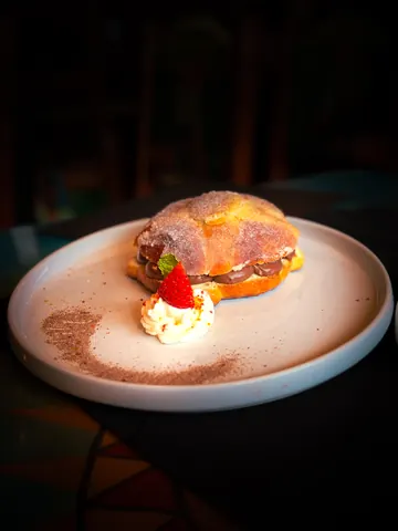 A pan de muerto split and filled with chocolate cream, plated with whipped cream, a strawberry and a dusting of cocoa